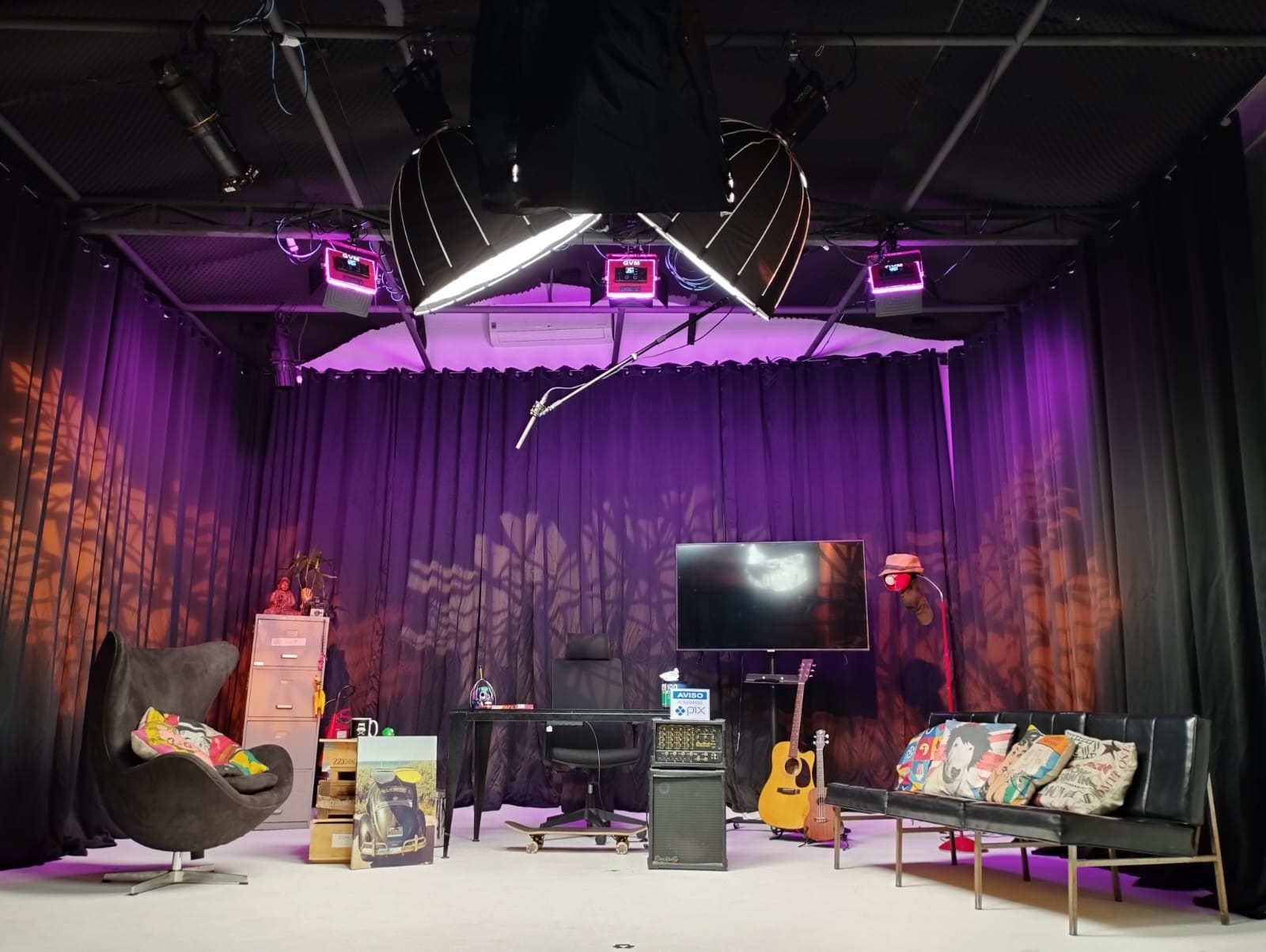 Um estúdio de gravação decorado com temática aconchegante e iluminação em tons de roxo e laranja. Há uma poltrona preta à esquerda com almofadas coloridas