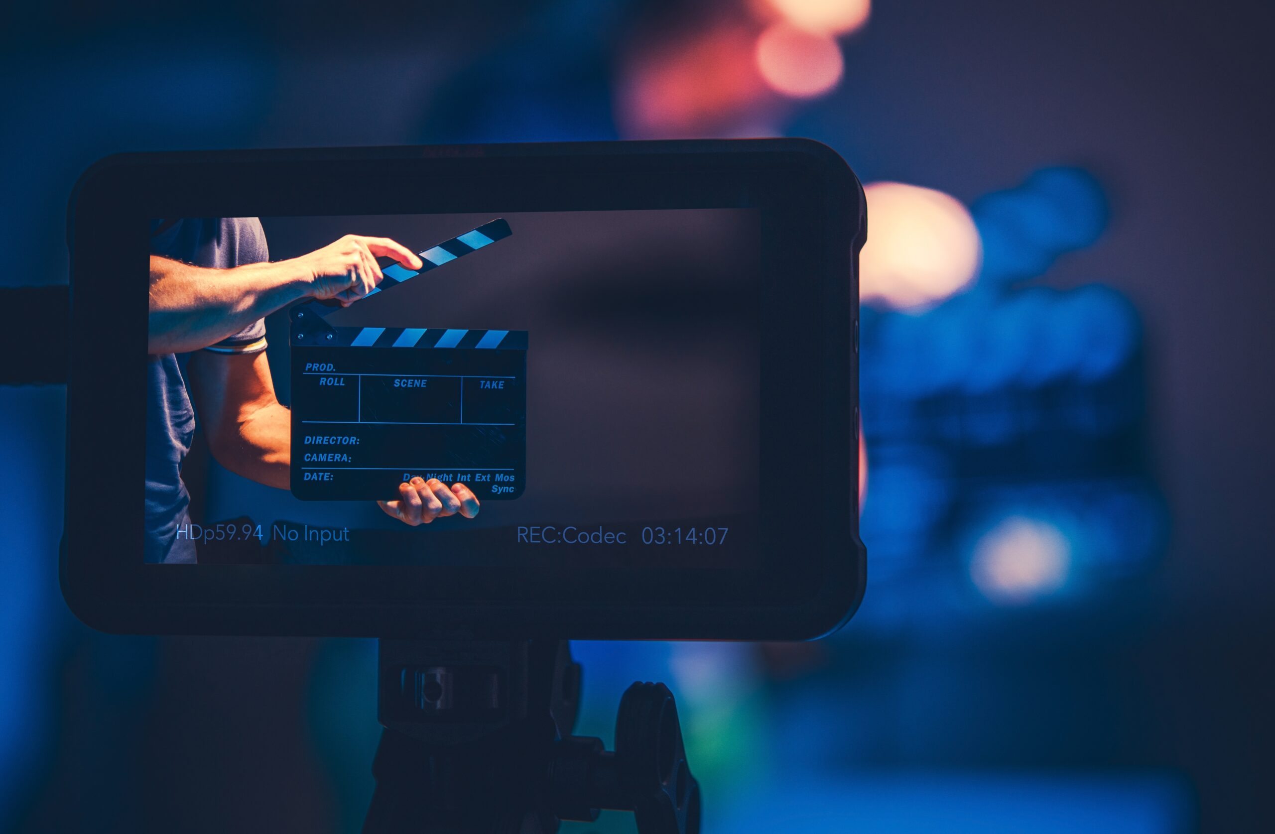 Um close-up de um monitor de câmera de vídeo mostrando uma claquete sendo usada no set de gravação. A cena está iluminada com luzes azuis e roxas, criando um ambiente de estúdio de filmagem. A claquete é segurada por uma pessoa e está pronta para marcar o início de uma tomada.
